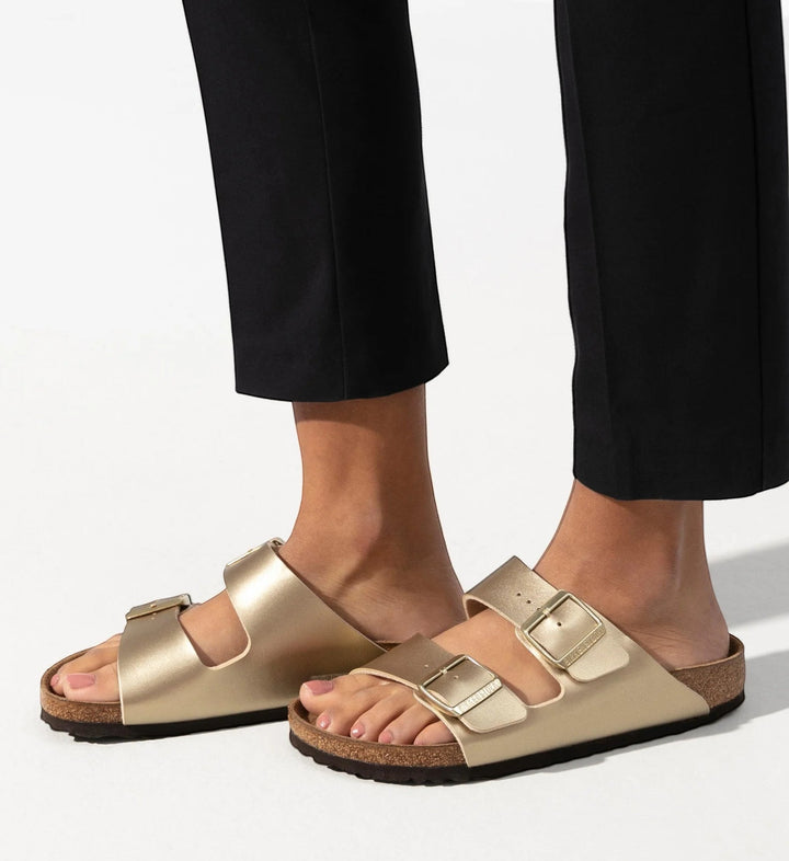 House of Florian Arizona Birko-Flor metallic gold double-strap sandals with cork footbed on a person wearing black cropped pants.