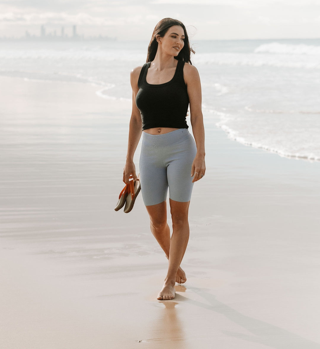 House of Florian woman in black tank top and light blue ribbed biker shorts walking on beach holding sandals.