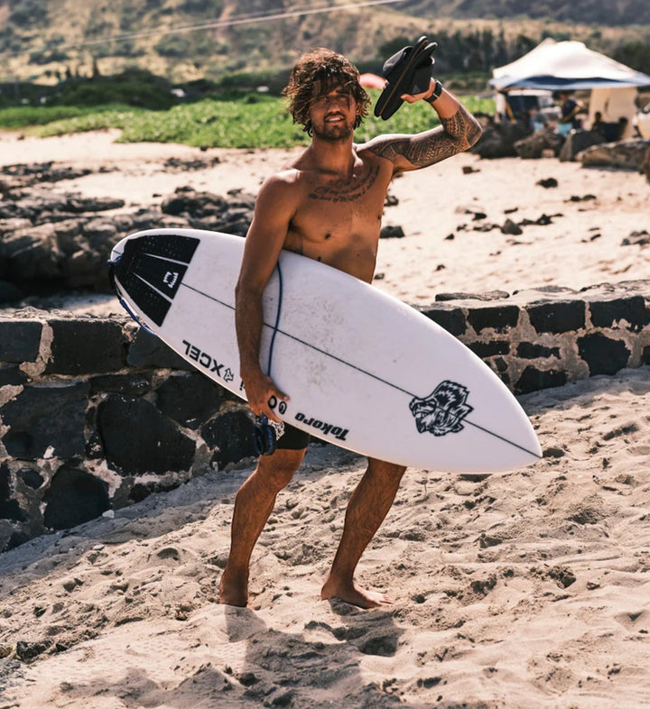 House of Florian Ulele Lanai Lava Rock surfboard with black and white graphics, held by a surfer on a sandy beach.